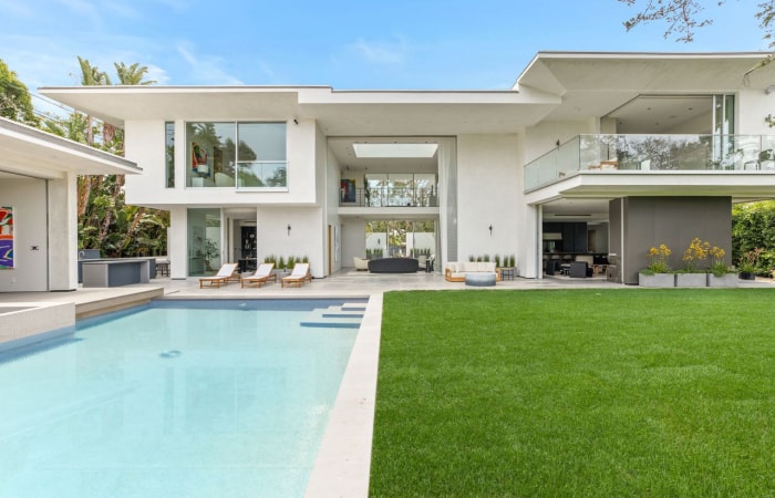 A sleek two-story white house features expansive glass windows and a balcony with glass railing, seamlessly connecting the open-concept expanse. A clear blue pool dominates the left side of the backyard, while lush green grass graces the right.