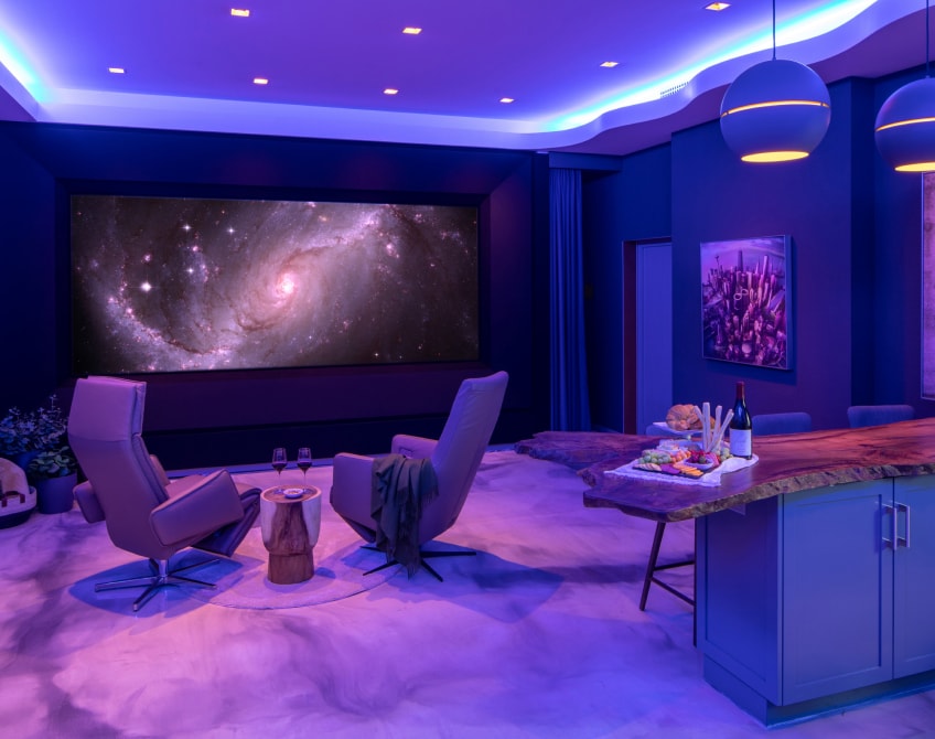 The Global Wave Showroom radiates in tranquil purple light, featuring a mesmerizing galaxy view on its theater screen, accompanied by opulent chairs and a carefully crafted wooden island adorned with delicious cuisine and wine.