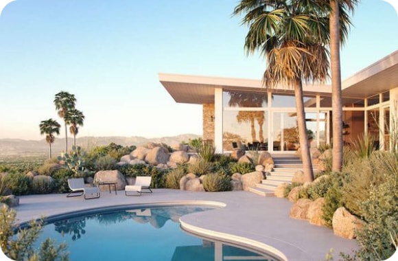 The luxury home's desert-themed backyard features distant mountains, palm trees, rocks, and green bushes, surrounding a curved blue pool.