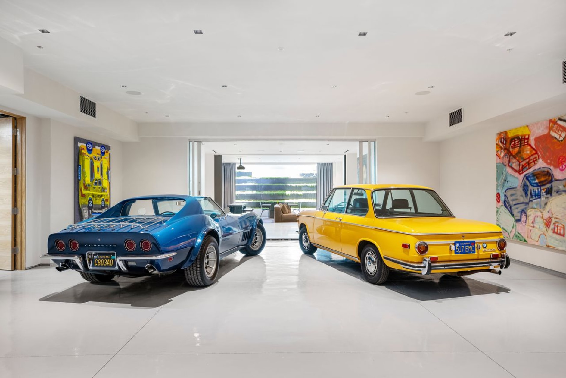 Impeccably restored vintage cars, one in vibrant yellow and the other in striking blue, command attention in a spacious white room, surrounded by vibrant paintings that echo the automotive theme.