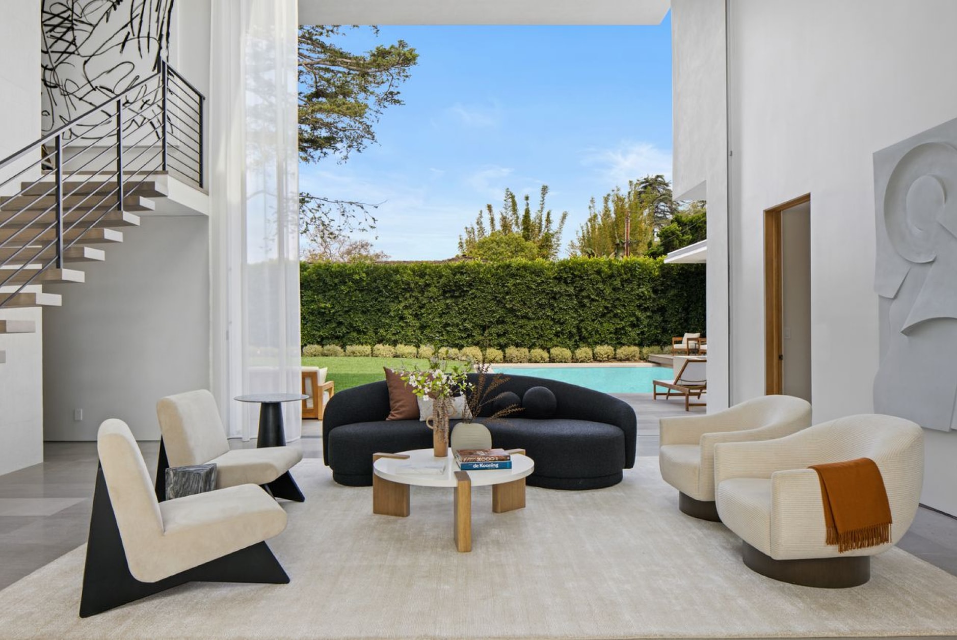 In a luxurious living room, a black couch is accompanied by four matching white chairs on a white rug. The back wall opens to a backyard with a clear blue pool, manicured grass, and a seamless hedge enclosure.