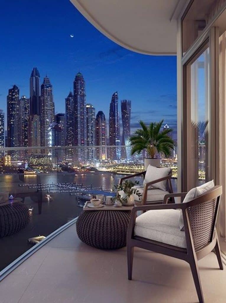 The balcony hosts chic lounge chairs paired with a coffee table, featuring white cushions and woven wicker frames, while the glass wall frames the enchanting illuminated New York City skyline at night.