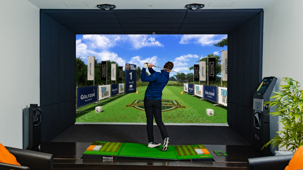 A man stands mid swing in front of a golf simulator screen.